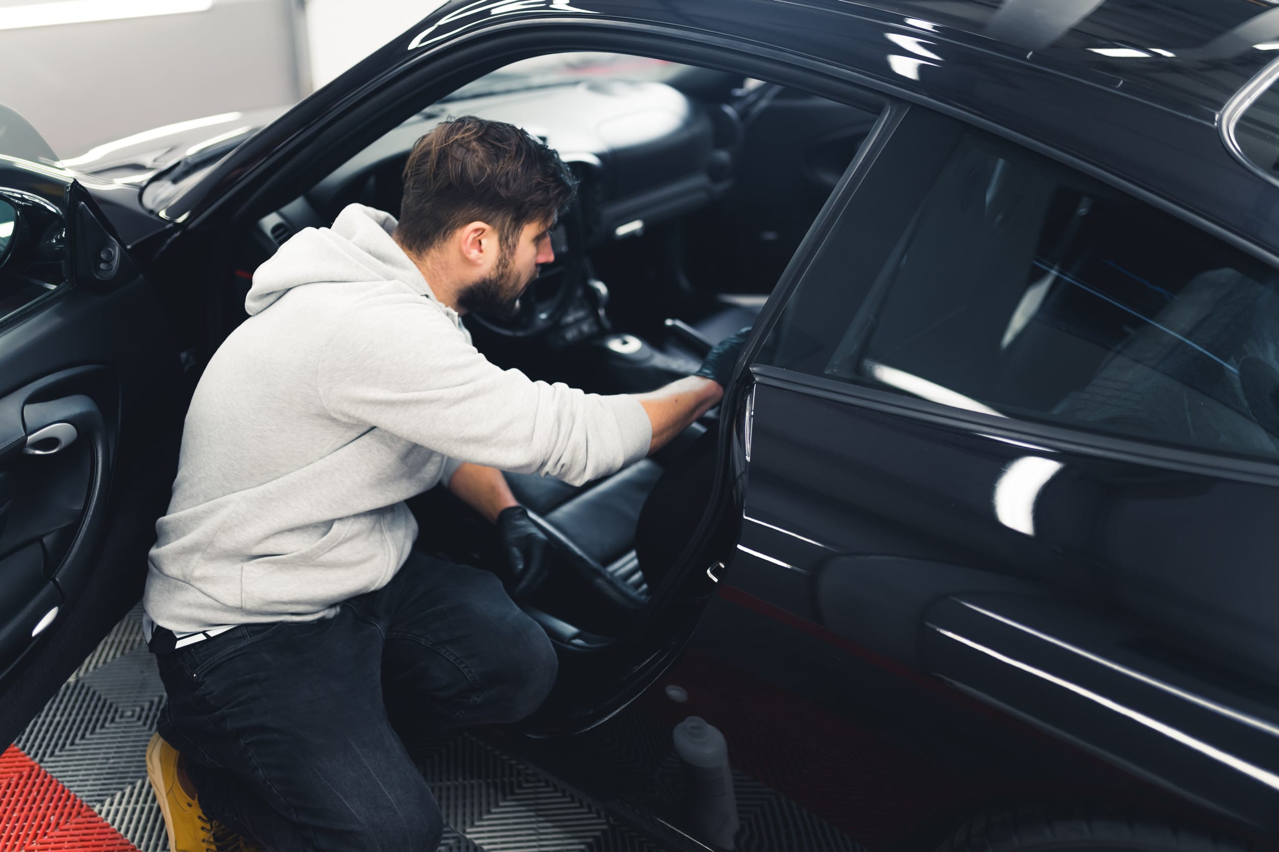 Man kneeling next to black car with open front door and cleaning leather car seats with cotton pad. Car detailing process.
